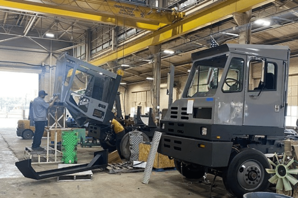 Yard Truck Specialist in New Boston, TX by Renew Truck. Inside a factory, workers assemble gray industrial vehicles. One vehicle cab is mounted on a chassis at right, while another cab is being lifted and installed using a hoist amid tools, parts, and overhead cranes.
